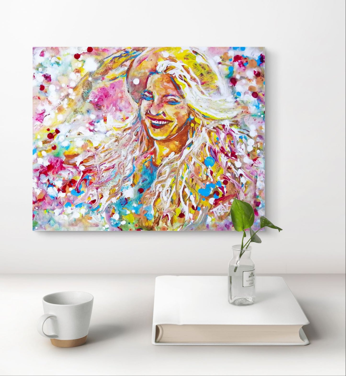 A colorful abstract portrait canvas print hanging on a white wall above a white table, with a white mug and a clear vase holding a green stem on a white book. Sun in the Hair original abstract feminine portrait fluid acrylic painting depicting a smiling woman in a straw hat with pink and yellow summer light by Diana Dimova TRAXI ART.