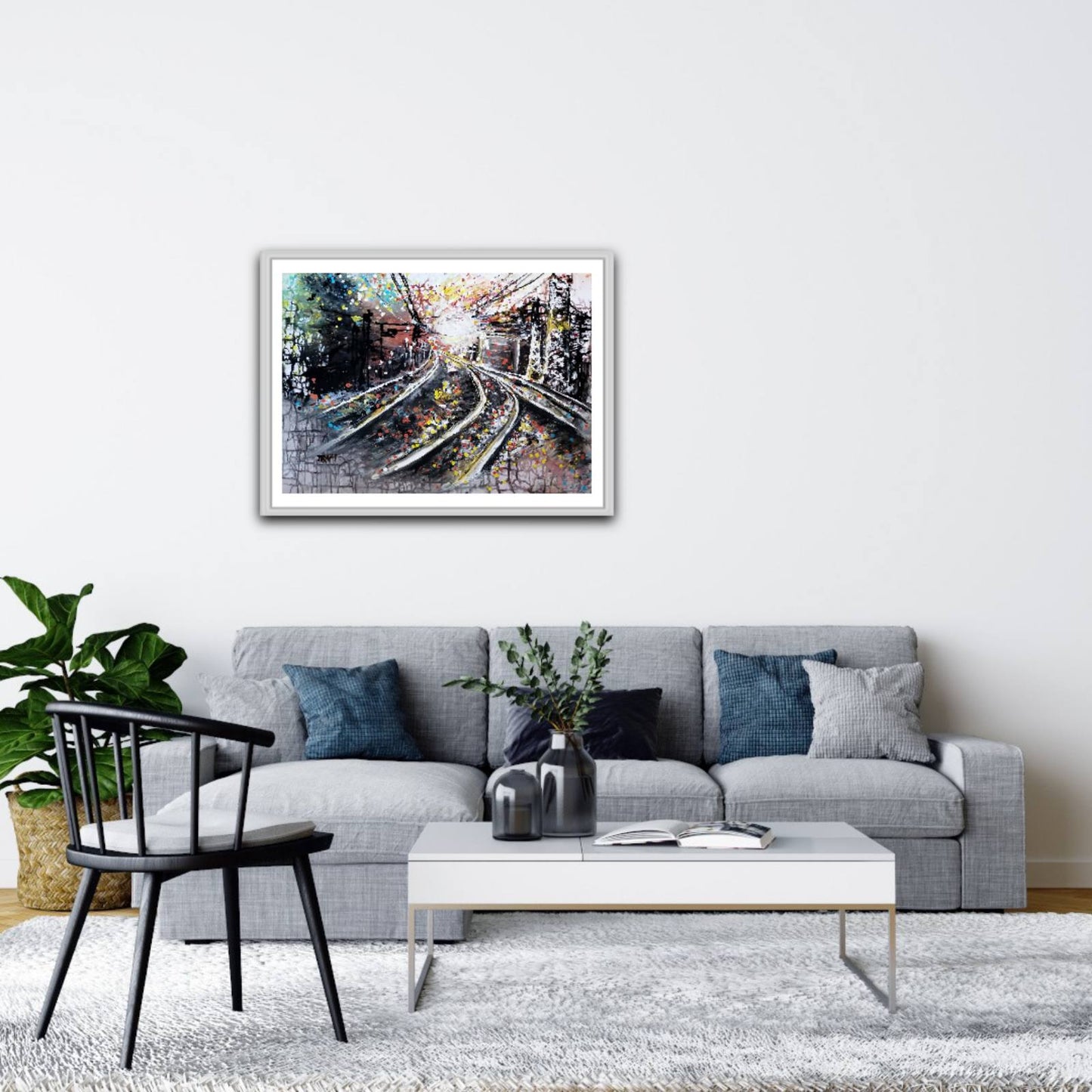 A framed art print depicting a detailed, colorful view of railway tracks is displayed on a white wall above a modern gray sofa in a styled living room setting. The Call of the Rails limited edition premium horizontal canvas print with minimalist black, brown, and white abstract railway lines by Diana Dimova TRAXI ART.
