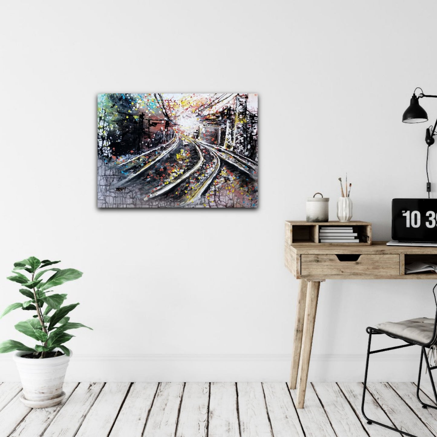 A rectangular abstract canvas print depicting intersecting train tracks, displayed on a white wall in a modern, minimalist room setting with a wooden desk, black lamp, and a potted green plant. The Call of the Rails original abstract urban landscape fluid acrylic painting with minimalist black, brown, and white tones representing railway lines by Diana Dimova TRAXI ART.