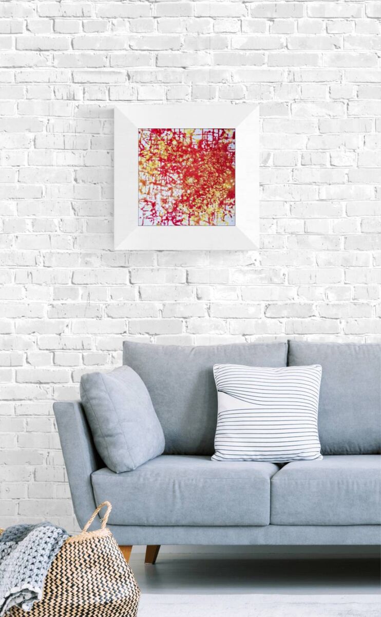 The Eye of Dawn original acrylic pouring abstract painting in red and gold by Diana Dimova TRAXI ART. Multi-award-winning 2022 artwork. A square framed abstract art print with red, orange, and yellow paint splatters hangs on a white brick wall above a light blue-gray sofa with decorative pillows.