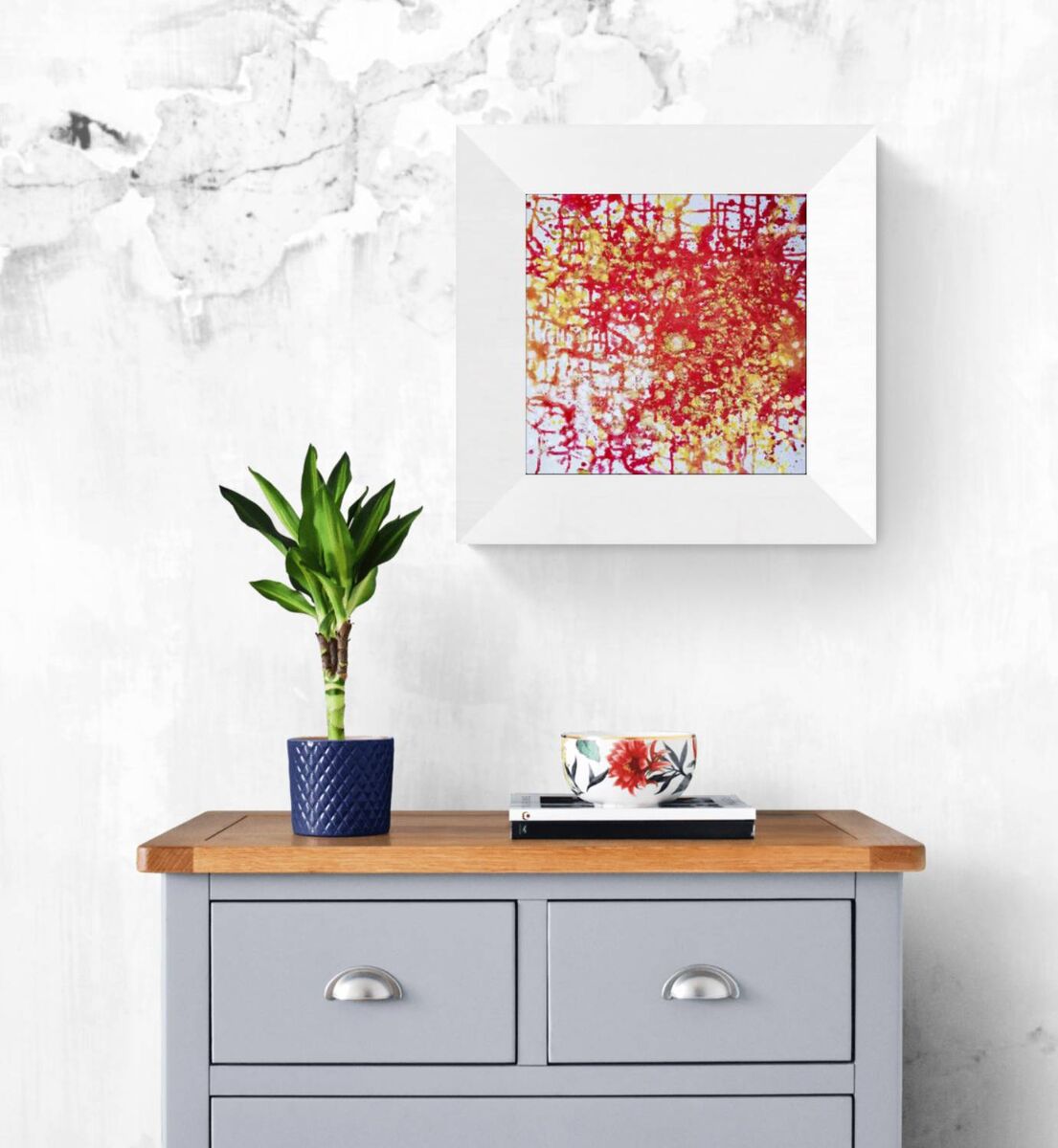 The Eye of Dawn original acrylic pouring abstract painting in red and gold by Diana Dimova TRAXI ART. Multi-award-winning 2022 artwork. A framed abstract art print with red, yellow, and white brushstrokes hanging on a textured white wall above a gray wooden cabinet with a plant and a small decorative bowl.
