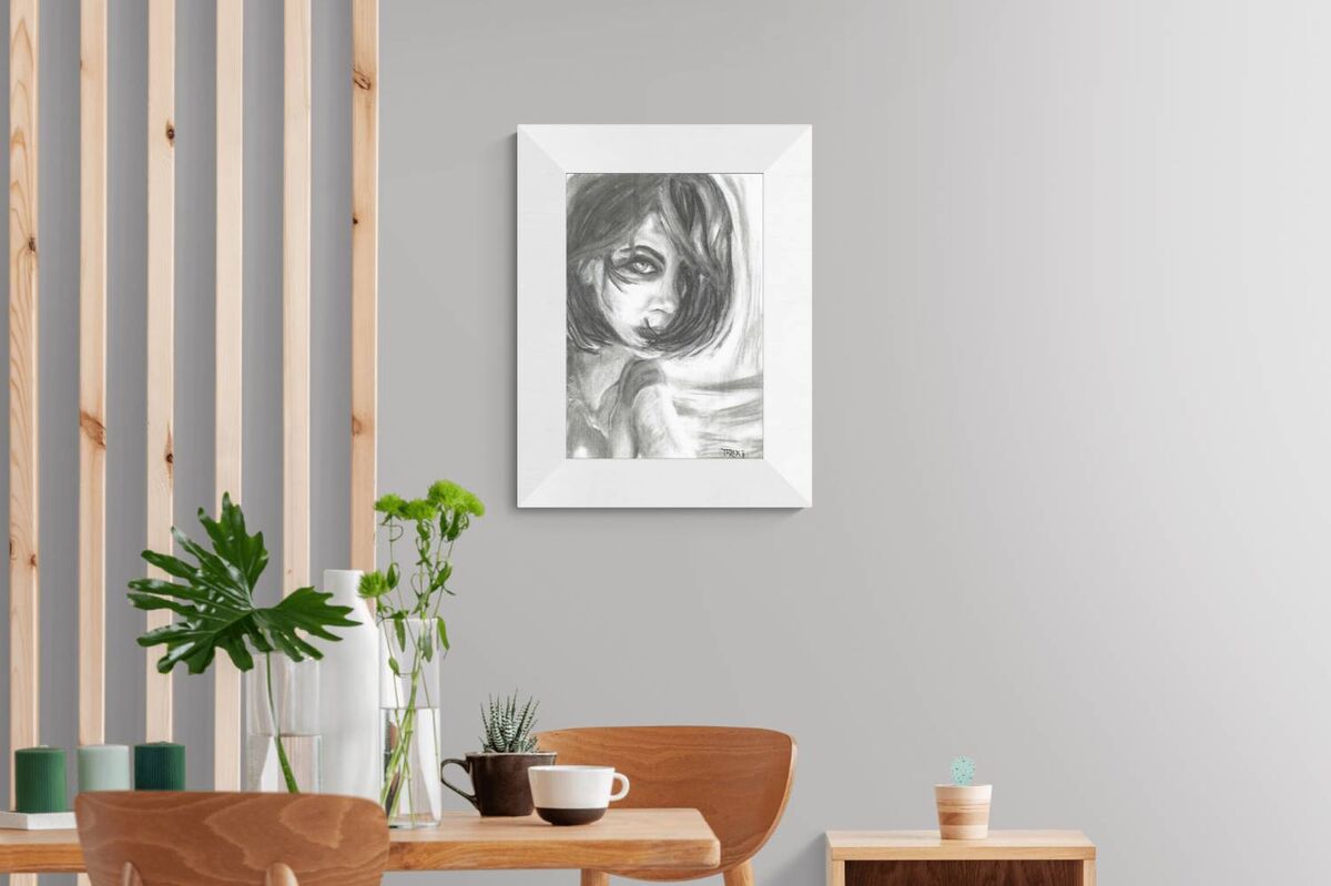 A framed black and white sketch portrait of a young girl hanging on a light gray wall in a modern, minimalist room with wooden furniture and plants.The Gaze Says It All original monochromatic charcoal portrait of a woman looking directly forward with a fearless gaze by Diana Dimova TRAXI ART.