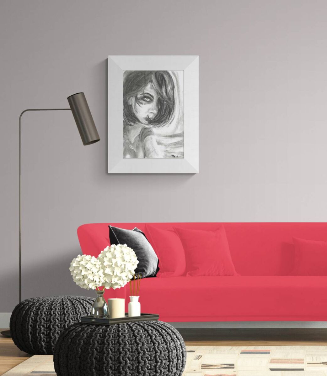 A framed black and white sketch of a girl's face hanging on a light gray wall above a bright red sofa in a modern living room setting. The Gaze Says It All original monochromatic charcoal portrait of a woman looking directly forward with a fearless gaze by Diana Dimova TRAXI ART.
