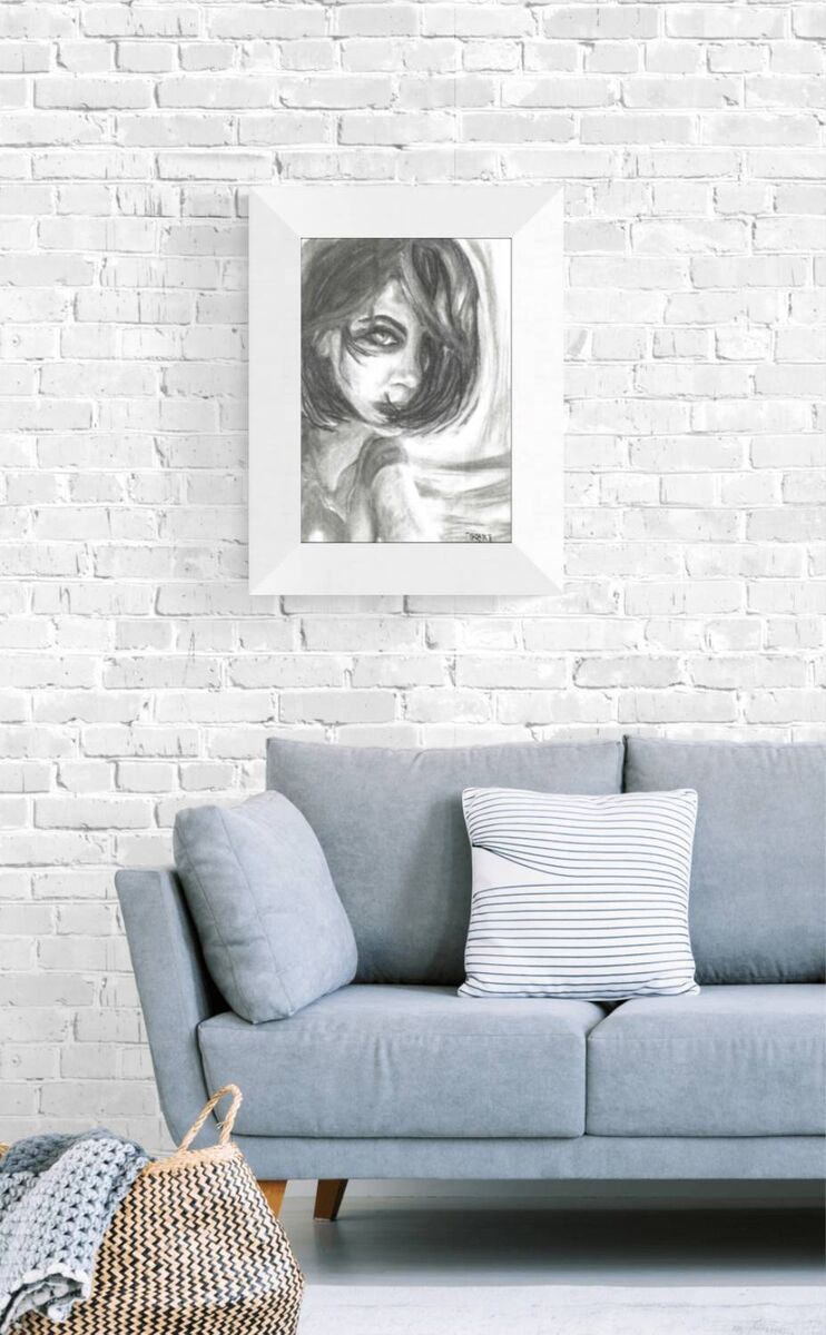 A framed black and white sketch portrait of a woman hanging on a white brick wall above a light blue sofa with pillows. The Gaze Says It All original monochromatic charcoal portrait of a woman looking directly forward with a fearless gaze by Diana Dimova TRAXI ART.