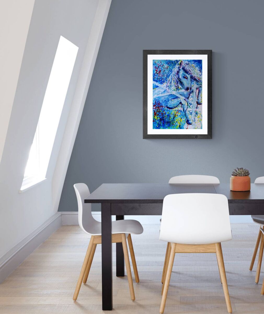 A framed abstract painting of a horse in vibrant blue, white, and green tones, displayed on a gray wall above a modern dining table and chairs. White Horse and Blue Freedom limited edition premium vertical canvas print depicting a symbolic white horse spirit with blue and red accents by Diana Dimova TRAXI ART.