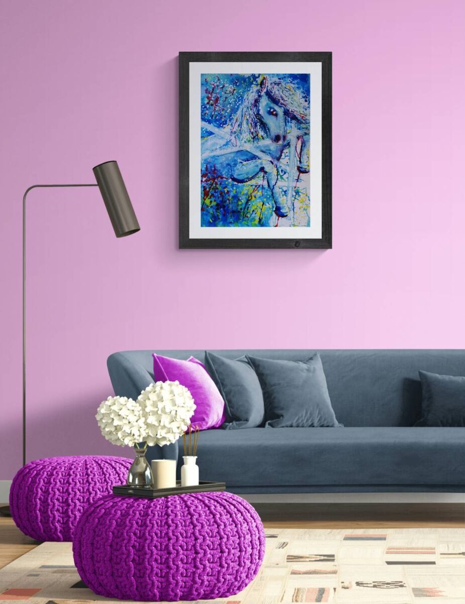 A framed abstract painting of a horse in vibrant blue, purple, and green tones, displayed on a light purple wall above a modern gray sofa with decorative pillows and a purple knit pouf in the foreground. White Horse and Blue Freedom original symbolic fluid acrylic painting with deep blue, white, and red accents by Diana Dimova TRAXI ART.