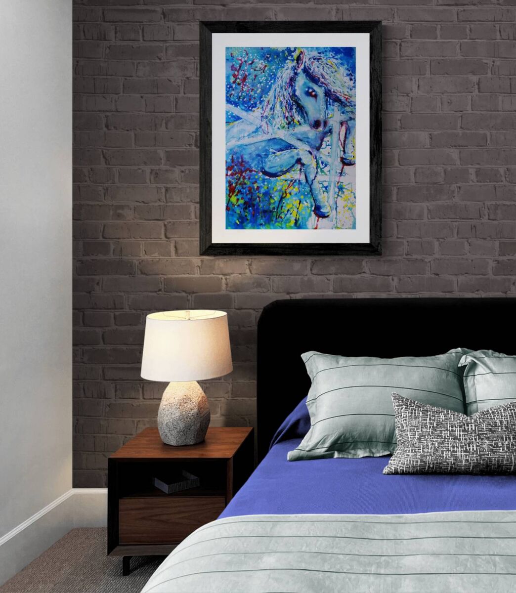 A framed abstract painting of a horse in vibrant blue, white, and red tones, displayed on a dark gray brick wall above a bed with blue bedding and a wooden nightstand. White Horse and Blue Freedom original symbolic fluid acrylic painting with deep blue, white, and red accents by Diana Dimova TRAXI ART.