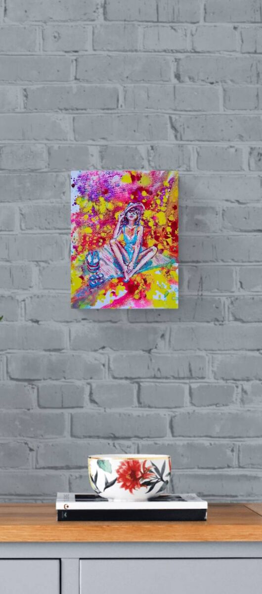 Wonderful Sunny Mood original acrylic pouring painting by Diana Dimova TRAXI ART, capturing an aura of light and summer energy. A colorful abstract painting of a seated figure in blue, pink, yellow, and red tones, displayed on a gray brick wall above a wooden surface with a white cup and books.