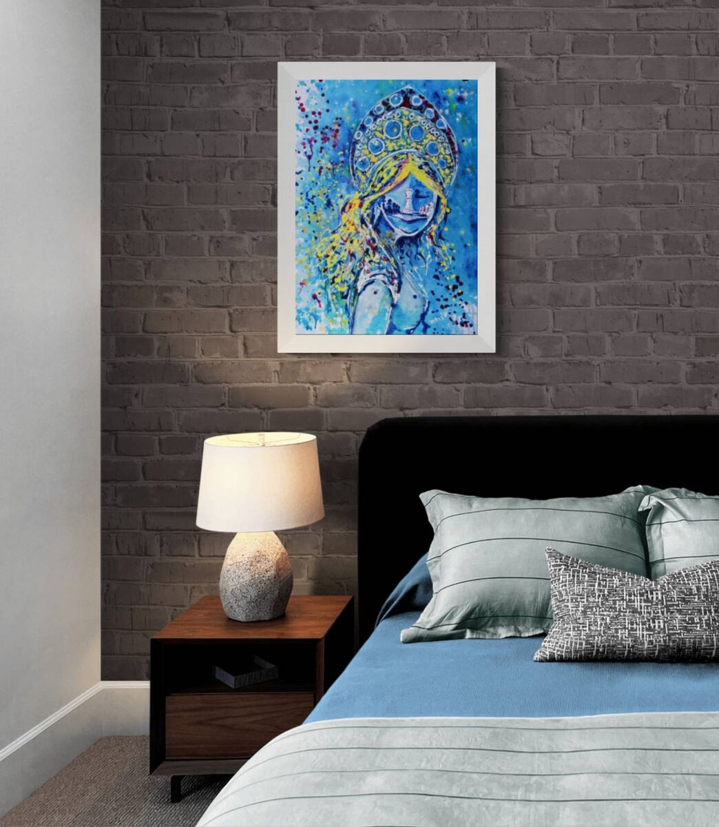 Original spiritual abstract painting titled White Queen’s Move by Diana Dimova TRAXI, featuring a cosmic goddess and chess symbolism in luminous blue and white tones. A framed abstract painting of a woman in a blue and yellow headdress, displayed on a dark gray brick wall above a bed with blue bedding and a wooden nightstand with a lamp.