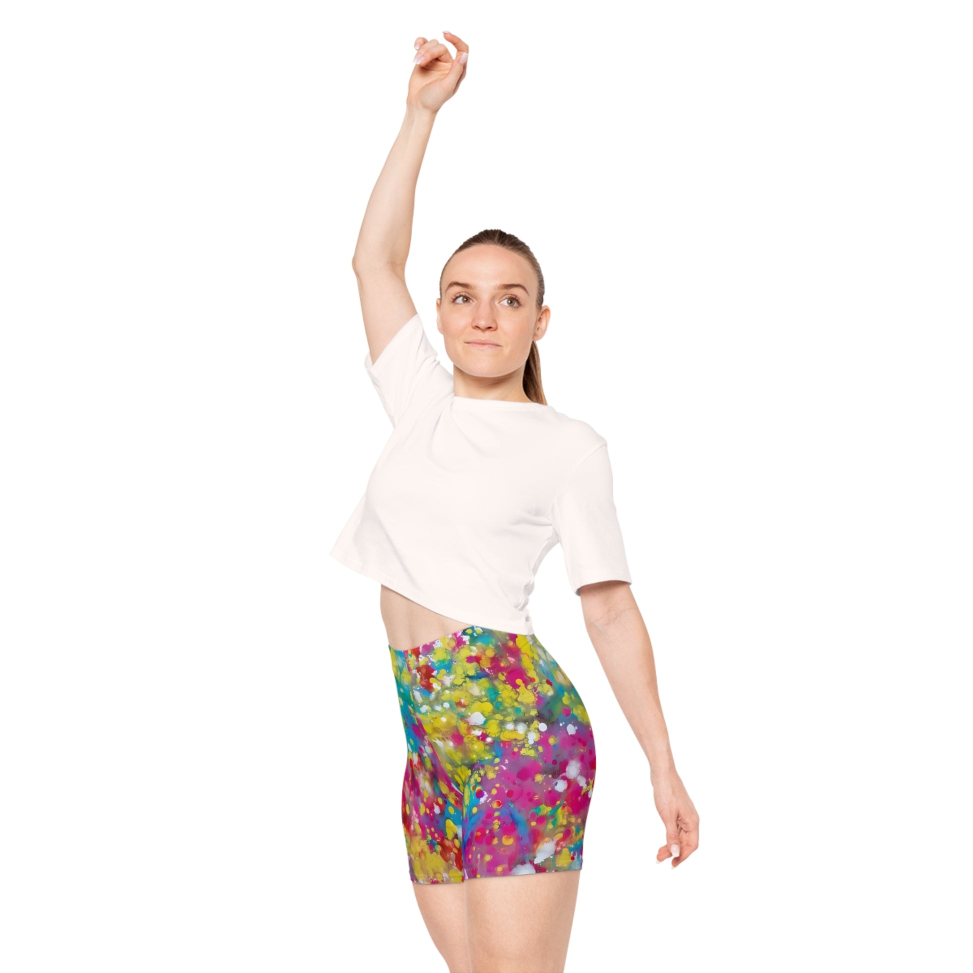 Women's summer biker shorts of happiness featuring abstract wearable art inspired by the Paintings of Happiness series by Diana Dimova TRAXI. A woman modeling a white short-sleeve crop top and high-waisted athletic mini skirt with a vibrant multicolor abstract paint splatter pattern, set against a plain white background.